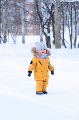 Fototapeta na wymiar Portrait of a toddler 15-20 months old in yellow warm clothes standing on a path and smiling in the middle of a winter park during a snowfall