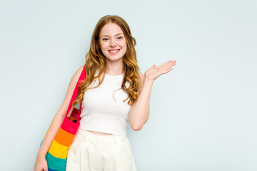 Young caucasian woman holding LGTBI bag isolated on blue background showing a copy space on a palm and holding another hand on waist.