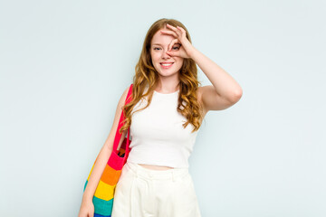 Young caucasian woman holding LGTBI bag isolated on blue background excited keeping ok gesture on eye.