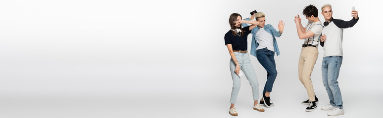 young woman showing victory sign and dancing near friends taking photo on smartphones on grey background, banner.