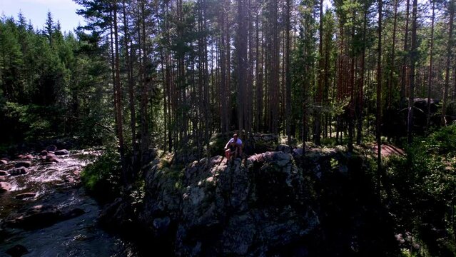 Arc shot of a father and daughter sitting in the Swedish wilderness on a gray cliff enjoying nature. River full of boulders. The sun shines through the evergreen pine trees. Family time in summer