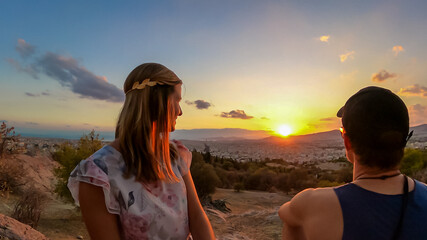 Young couple  watching sunset over city of Athens seen from Filopappou Hill (hill of muses), Athens, Attica, Greece, Europe. Athens cityscape and Aegean sea. Beautiful sunset point with aerial view