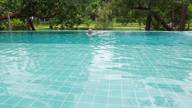 A woman sits on the edge of the pool and swims against the backdrop of tropical vegetation in a hotel or villa.