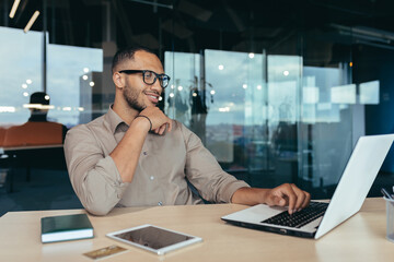 Happy and successful Indian man working in modern office building, programmer in glasses using laptop to write code, businessman in shirt smiling and happy .