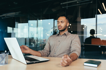 Successful businessman working inside modern office building, man meditating with eyes closed in lotus position sitting on chair.
