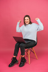 Ecstatic victorious young woman is sitting on a chair with laptop on her knees.