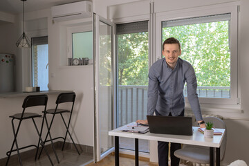 Portrait of a smiling young businessman leaning on his desk in a home office working online with a laptop. 