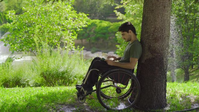 Adventurous physically disabled teenager reading a book outdoors.
Disabled teenager leaning his wheelchair against a tree and reading a book.
