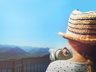 blonde girl with straw hat stands at public telescope in front of swiss mountain scene and clear blue sky. holiday and look into future concept postcard motive. selective focus on telescope eyepiece