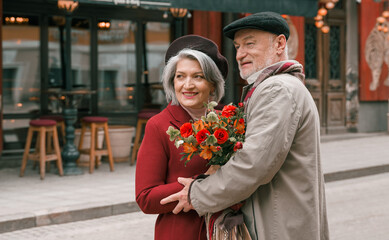 Elderly senior love couple. Old retired man woman together on romantic date.Aged husband wife walking on city street with flowers.Stylish elder hugging people pensioner in red coat.Happy family years