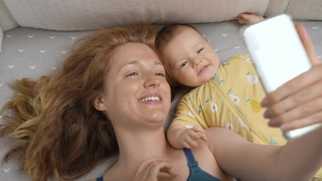 Young mother and her newborn baby making a selfie or video call in a bed. Technology, family, parenthood. Top view
