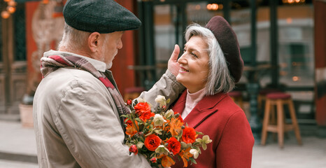 Elderly senior love couple. Old retired man woman together on romantic date.Aged husband wife walking on city street with flowers.Stylish elder hugging people pensioner in red coat.Happy family years