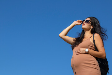 Heavily pregnant brunette woman in a beige dress, black sunglasses and a backpack touching her tummy and looking upwards into the distance on the clear blue sky background