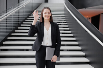 Fototapeta premium business woman in a formal suit with a laptop in the office of the business center stands on the steps and greets colleagues. Work in a corporation as a marketing financier or lawyer. a successful
