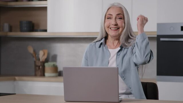 Happy middle aged senior grandma sit at home use laptop check email message read positive news get sale offer feel excitement make yes gesture celebrate success win online lottery career opportunity 