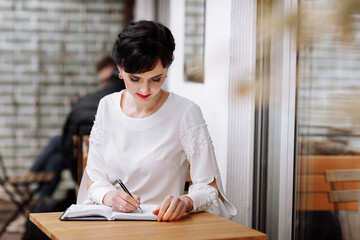 Serious focused smart businesswoman is writing notes on notepad, making agenda on personal organizer. Female in business suit sitting at table at cafe on terrace, work and event planning.