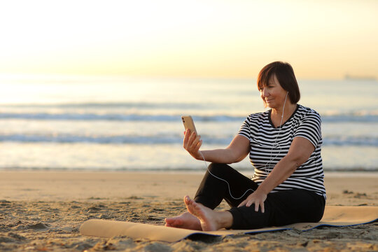 middle ages woman takes a break while sitting and relaxing on the yoga fitness mat and talking on smartphone with headphones on the beach by the sea. outdoor workout. Healthy, chill out lifestyle.