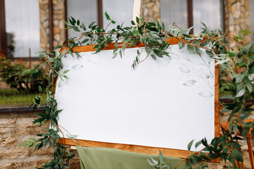 Wedding wooden board, mockup, invitation easel, with white space for an inscription, decorated with fresh greenery and green cloth. Wedding frame outdoors. Festive decor, copy space.