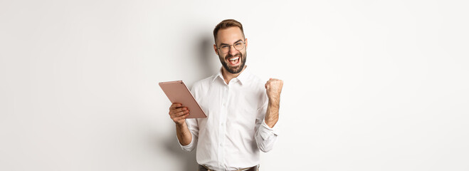 Successful businessman rejoicing on winning online, reading on digital tablet and making fist pump,...