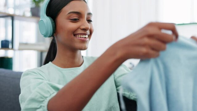 Happy woman, wireless headphones and cleaning while listening to music and folds clean laundry or washing while sitting on sofa at home. Indian female singing and streaming radio and having fun