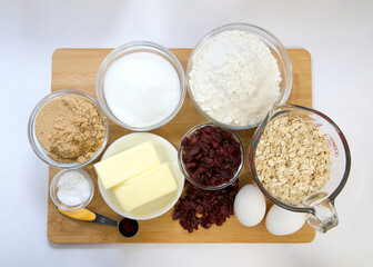 Top view flat lay of measured ingredients in bowls and cup for cranberry oatmeal cookies. On wood cutting board with parchment paper underneath.