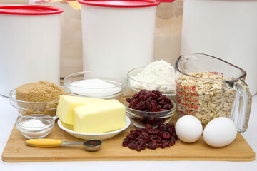 Measured ingredients in bowls and cup for cranberry oatmeal cookies. On wood cutting board with parchment paper underneath. Plastic canisters in background.