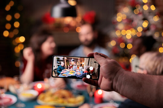 Family christmas celebration photo on smartphone screen, friends eating at festive dinner table on blurred background. Xmas holidays celebrating, hand holding mobile phone close up