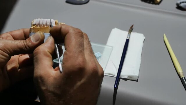 The hands of a dental technician model a removable ceramic denture using a sharp scalpel at a work table in an orthodontic dentistry workshop. Production and installation of dentures