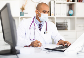 Doctor in protective medical mask sitting at workplace with computer in her office