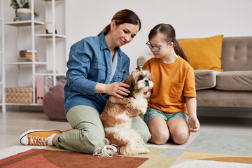 Full length portrait of mother and daughter with Down syndrome playing with dog on floor in cozy...