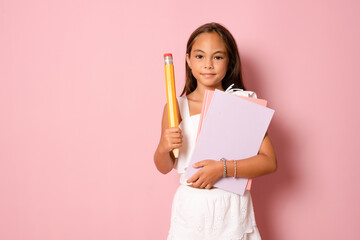 Cute pretty child girl with books on pink background holding a big pencil.