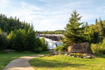 A path leads through a break in the trees towards Wawa Falls in the small town of Wawa, Ontario during an early evening sunset.