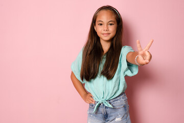 Happy little girl smiling showing V peace sign hand gesture on pink background. Peace to the world, stop the war