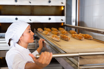Young Hispanic woman in white uniform working in bakery, pulling freshly baked baguettes out of...