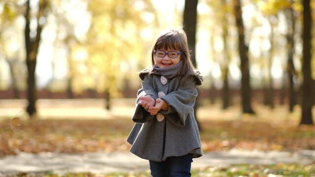 Happy cute brown-haired girl with down syndrome in fashionable coat and stylish eyeglasses tossing bright foliage up to the sky and laughing, kid enjoying time in warm autumn park, happy childhood
