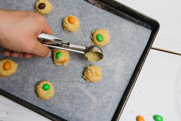 Halloween biscuits, sugar cookie dough with chocolate candies in colors orange and green, fun baking recipe for kids, male hand scooping raw cookie balls with scoop on baking sheet, white background - 532309505