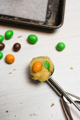 Sugar cookie dough with chocolate candies in Halloween treats colors orange and green, fun baking recipe for kids, scooping raw cookie balls with scoop on baking sheet on white wooden background - 532309563