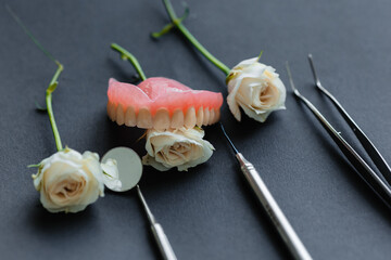 Forceps, tweezers, mirror, jaw, curettes and scalers lie in front of rose flowers on a black background. Aesthetics of dentistry.