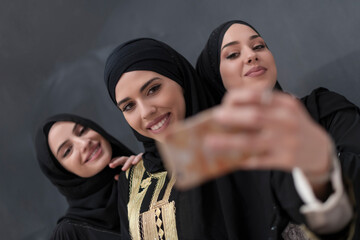 Group of young beautiful muslim women in fashionable dress with hijab using smartphone while taking selfie picture in front of black background