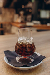 Ice coffee in a tall glass with whiskey. Cold summer drink on a dark napkin and white plate, light wooden table in café