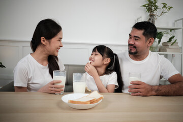 A Healthy Asian Thai family, a little daughter, and young parents drink fresh white milk in glass and bread joy together at a dining table in morning, wellness nutrition home breakfast meal lifestyle.
