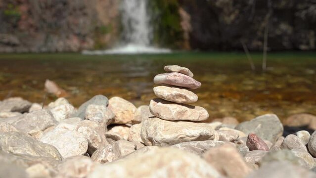 Woman hand builds pyramid of stones against backdrop of seething waterfall, reservoir wildlife. Cairn making wish. Superstition for luck, success. Traveler fun picnic, build figurines cobblestones