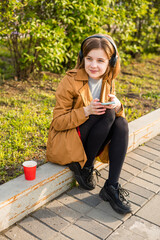 A young teenage girl sits on the street with a smartphone in her hand, headphones, listens to music. High quality photo