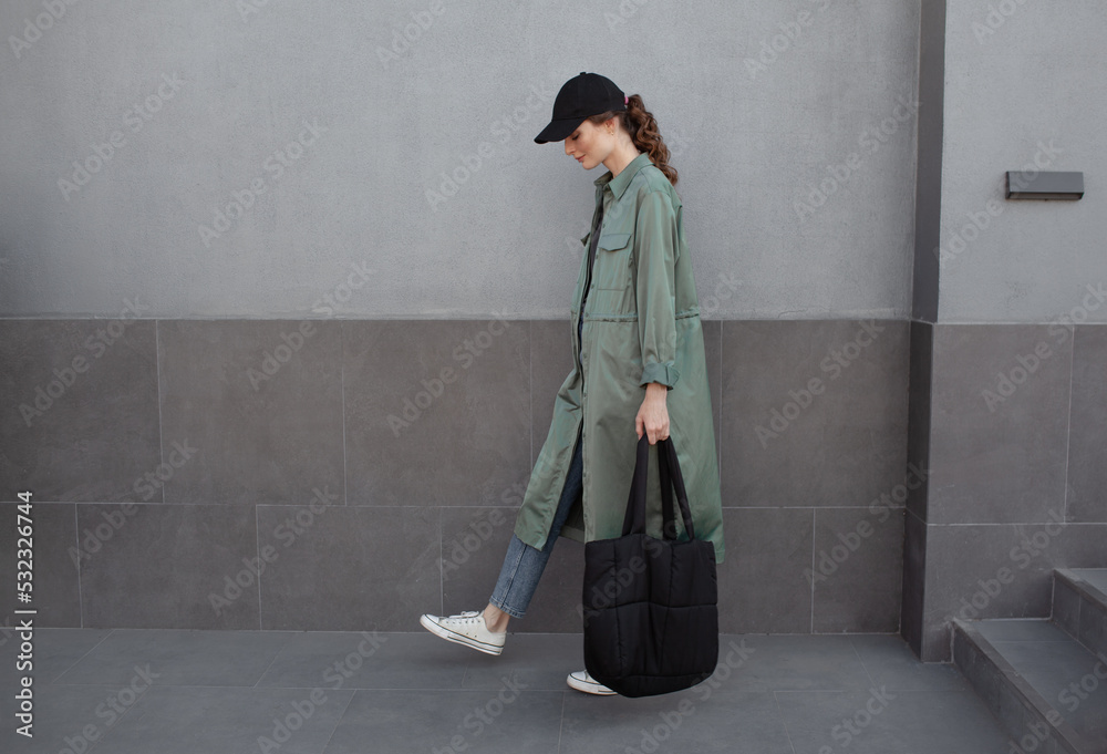 Wall mural A beautiful girl is wearing a grey t-shirt, cloth cap, coat, black bag and jeans posing against the background of a wall. fashion outfit for the city, minimalism urban style clothing