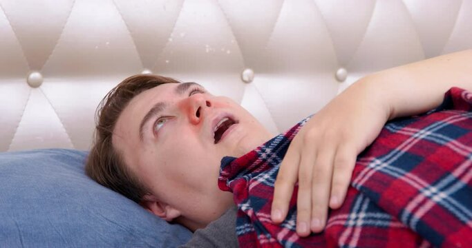 Adult man sneezes lying on bed with red checkered blanket. Brown-haired guy with red runny nose feels unwell deciding to rest recovering closeup