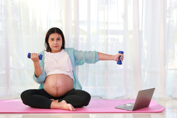 Happy young asian pregnant sit on pink mat lifting dumbbell and watch training video online from computer in living room at home. Expectant mother prepare for baby birth during pregnancy concept