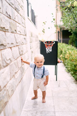 Little boy stands near a rack with a basketball basket against a stone wall. High quality photo