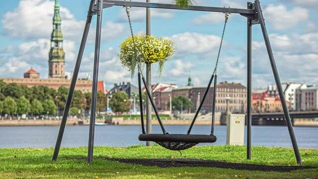 A swing along the banks of the Daugava River with St. Peter's Church steeple in city skyline and a cloudscape overhead - time lapse