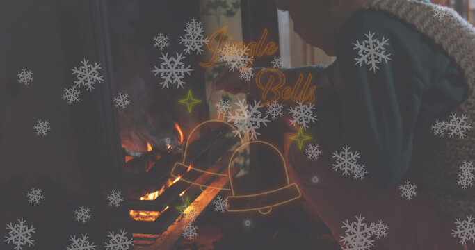 Image of jingle bells text in red neon, christmas bells and snowflakes over boy lighting fire