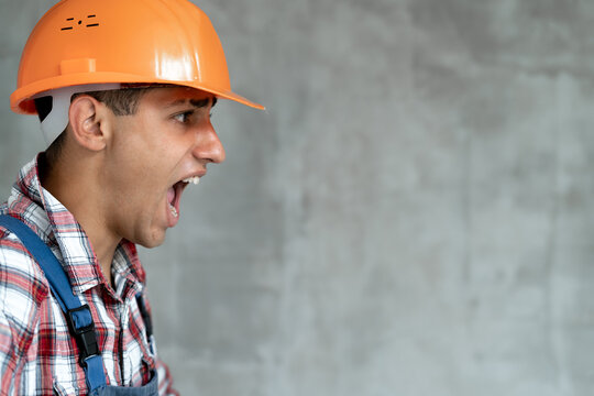 Young builder man wearing construction uniform and safety helmet over gray isolated background crazy and mad shouting with aggressive expression. Frustration